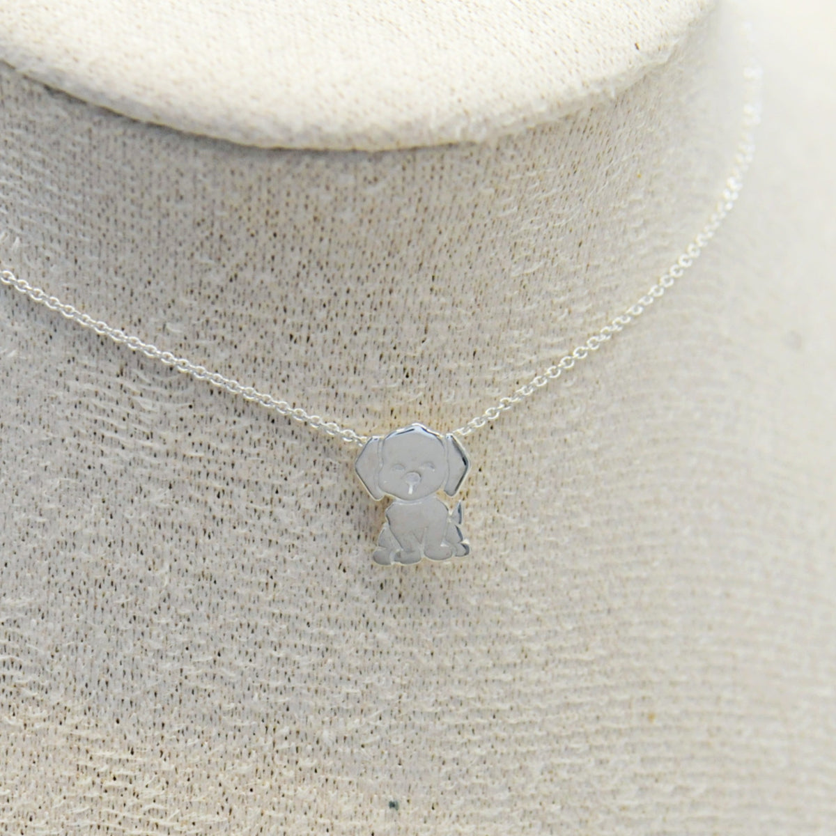 A delicate silver chain necklace with a small, stylized dog-shaped pendant, displayed on a textured beige fabric jewelry bust.
