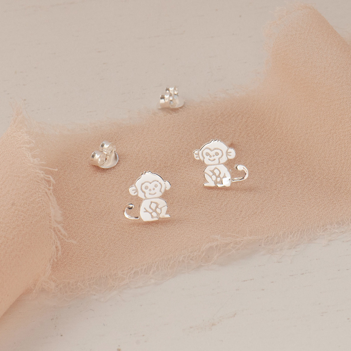 Silver monkey stud earrings displayed on a soft beige fabric surface against a light neutral background.