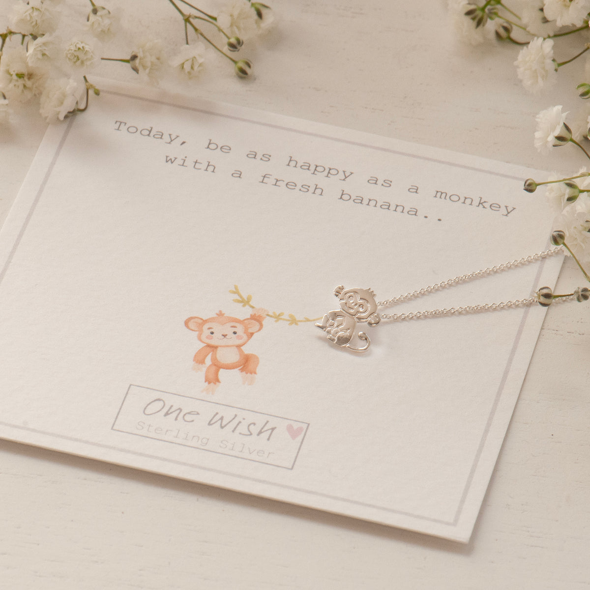 A silver monkey pendant necklace on a delicate chain, displayed on a white card with a monkey illustration and the text 'One Wish sterling silver.' The card is styled with white flowers on a light surface.