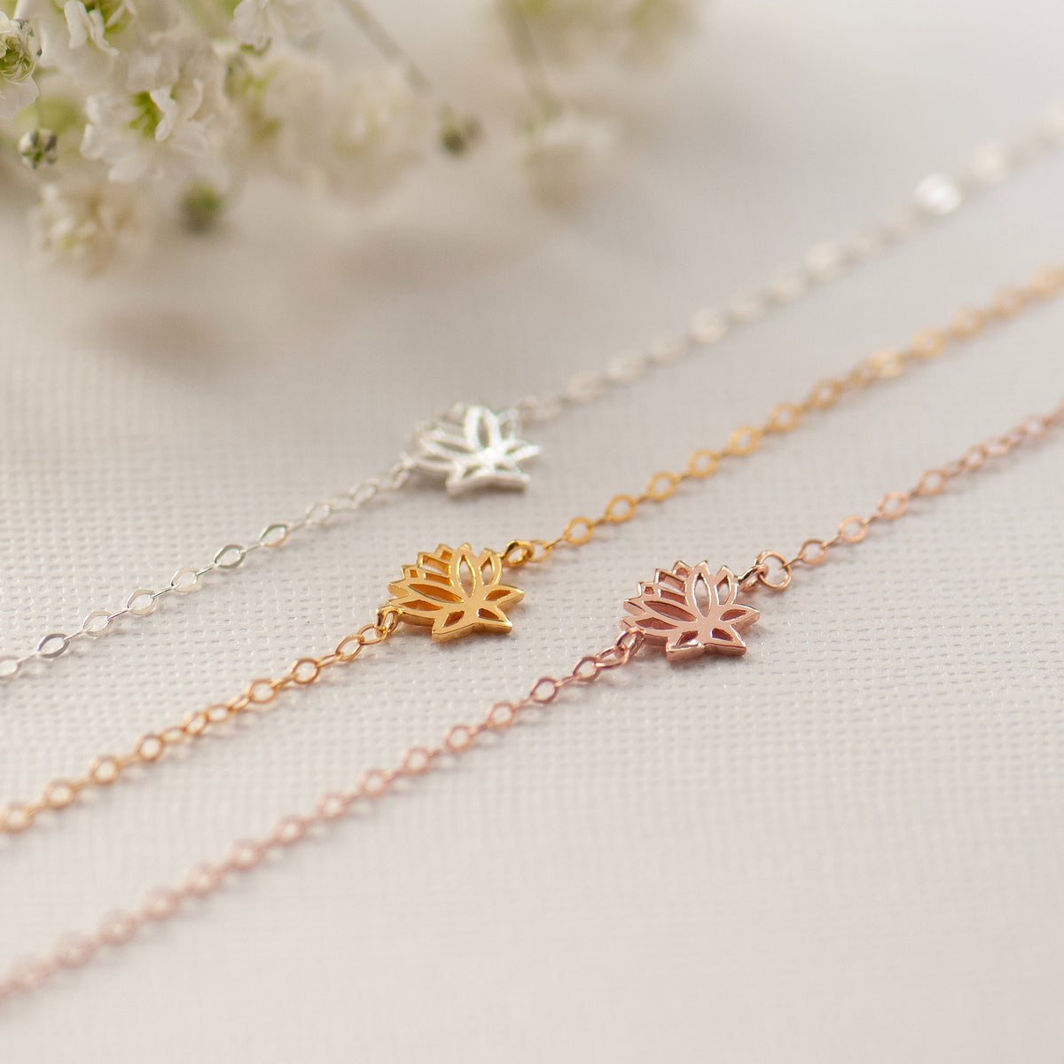Three delicate chain bracelets with small lotus flower charms in silver, gold, and rose gold, arranged diagonally on a textured cream fabric with soft white flowers in the background.