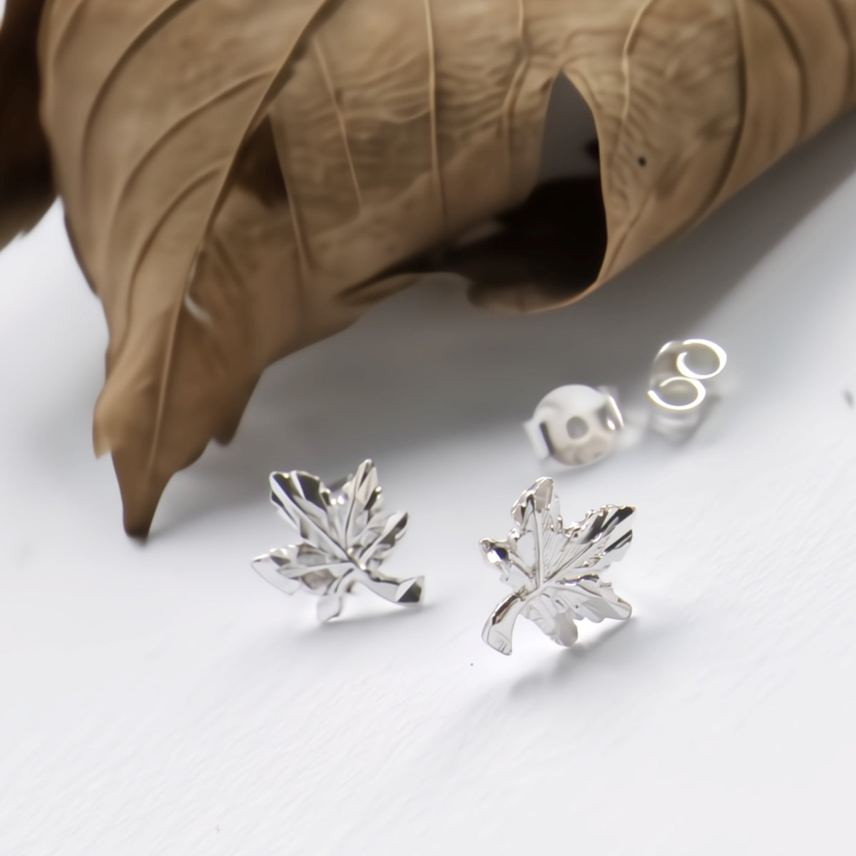 Silver maple leaf stud earrings displayed on a white surface next to a dried brown leaf, with one earring shown from the front and another showing the post backing.