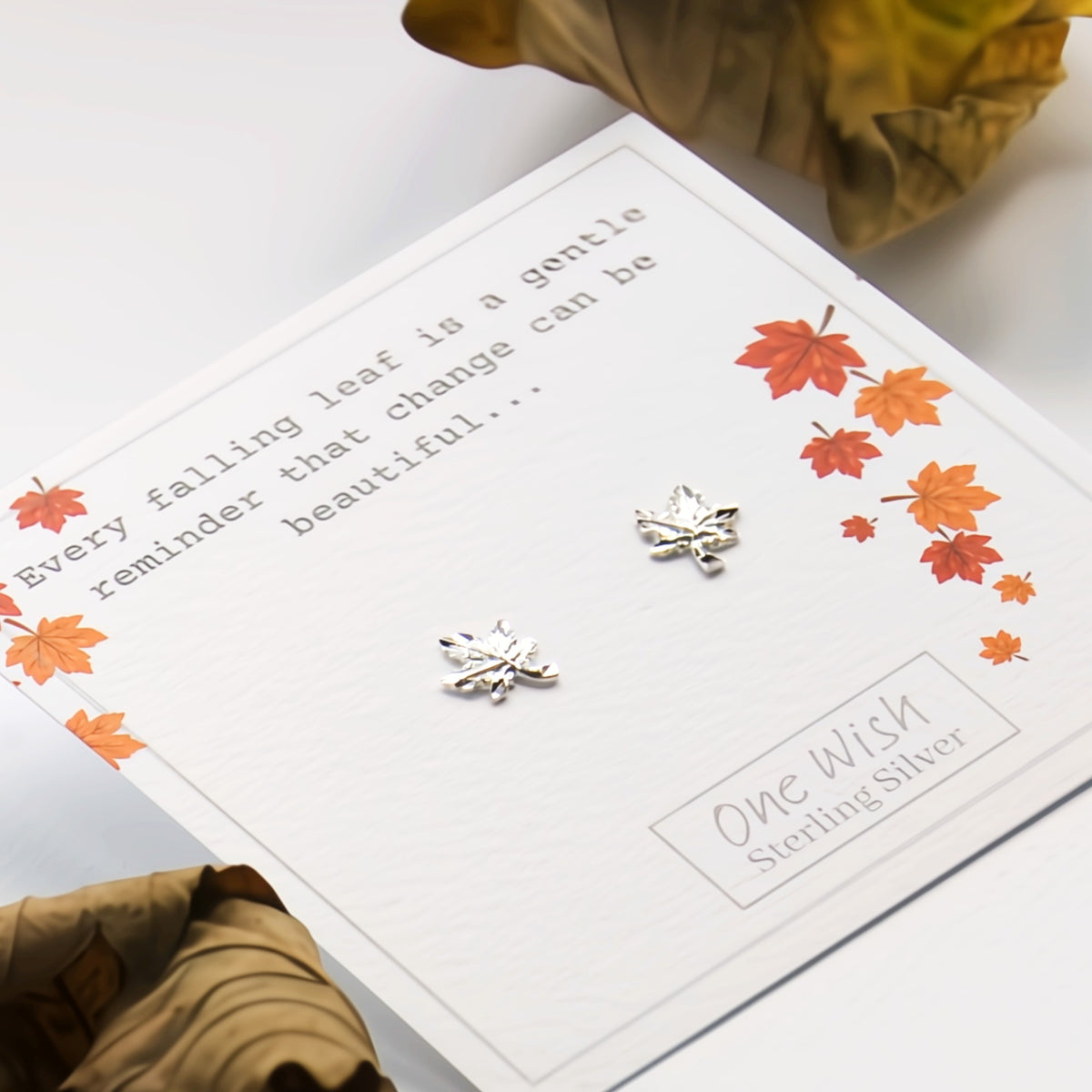 A pair of silver leaf stud earrings displayed on a white gift card with autumn leaf illustrations and the text 'One Wish Sterling Silver.' The card is photographed on a light surface with dried leaves as styling props.