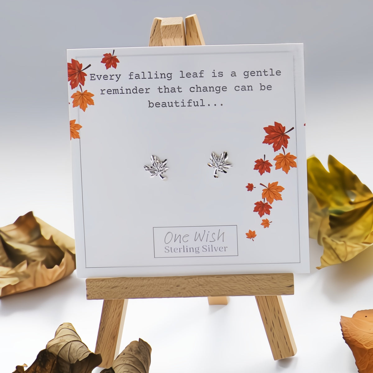 A pair of sterling silver leaf stud earrings displayed on a white card with autumn leaf illustrations and a quote, set on a small wooden easel with dried leaves around it.