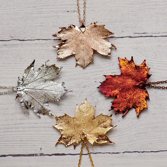 A collection of four Canadian maple leaf necklaces in different finishes: silver, gold, rose gold, and iridescent copper, displayed on a wooden surface.
