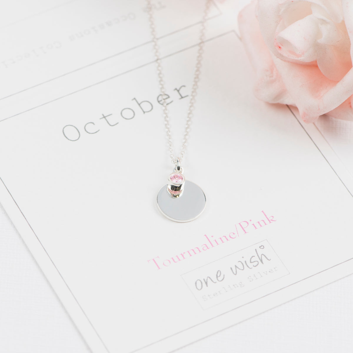 A silver necklace with a round disc pendant and a small pink tourmaline gemstone, displayed on a white card labeled 'October' with a pink rose in the corner.