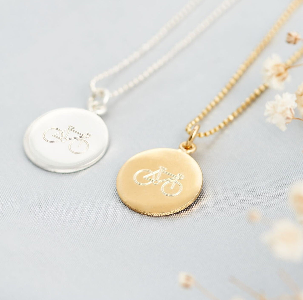 A sterling silver bicycle pendant necklace on a silver chain, with a gold-plated bicycle pendant alternative next to it, placed on a white surface with some dried flowers scattered around.