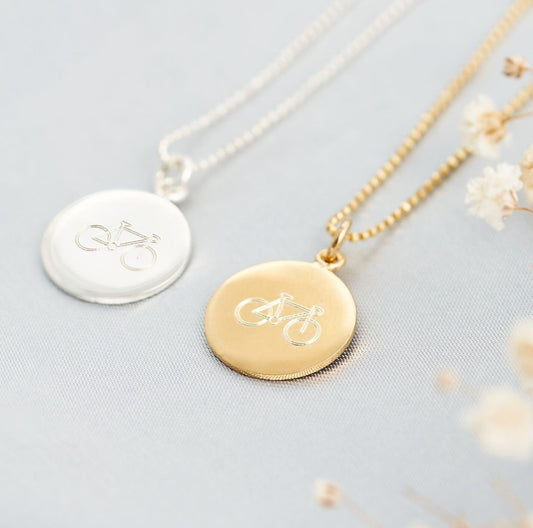 A sterling silver bicycle pendant necklace on a silver chain, with a gold-plated bicycle pendant alternative next to it, placed on a white surface with some dried flowers scattered around.