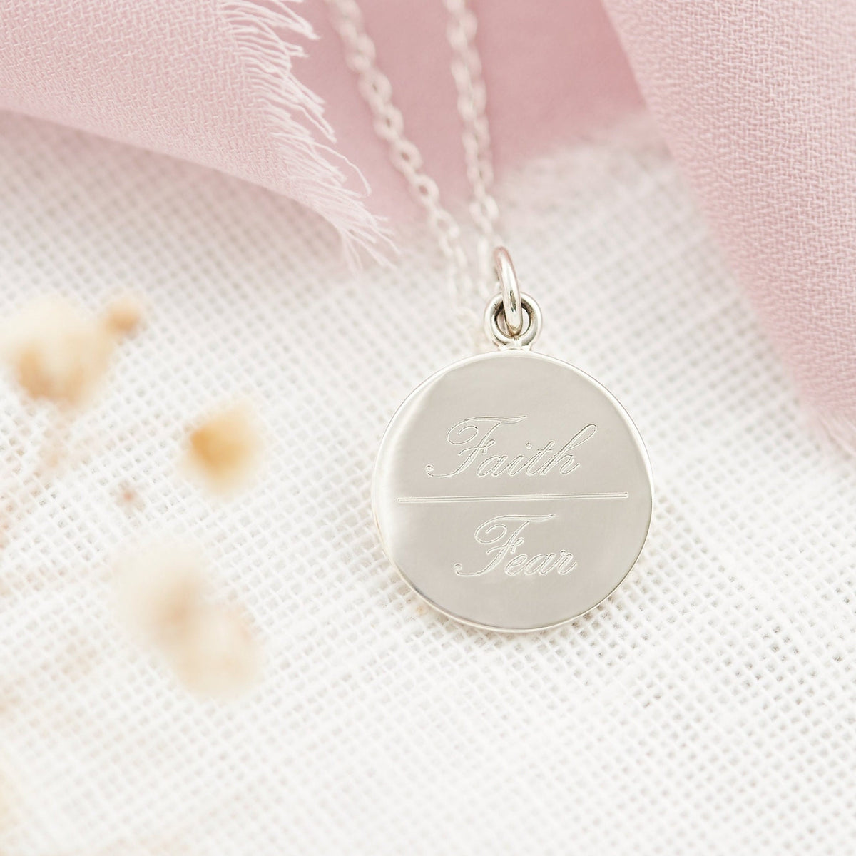 A sterling silver engraved pendant necklace with the inscription 'Faith Over Fear' displayed against a pink and white floral background.