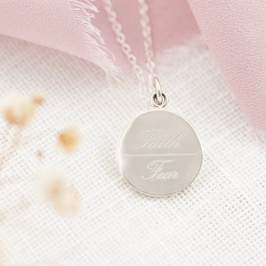 A sterling silver engraved pendant necklace with the inscription 'Faith Over Fear' displayed against a pink and white floral background.