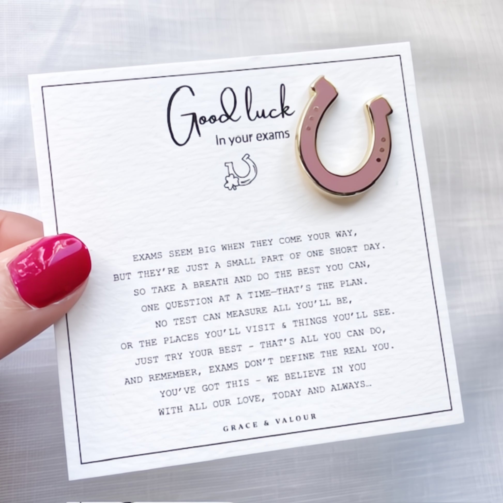 Pink and gold horseshoe charm on a card with a message of good luck for exams, held by a hand with red nail polish.