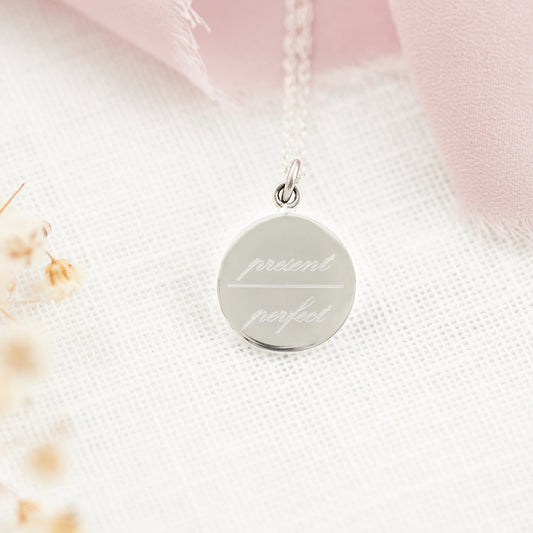 A silver round disc pendant necklace engraved with the words 'present perfect' on a delicate chain, resting on a textured white fabric with soft pink ribbon and dried flowers.