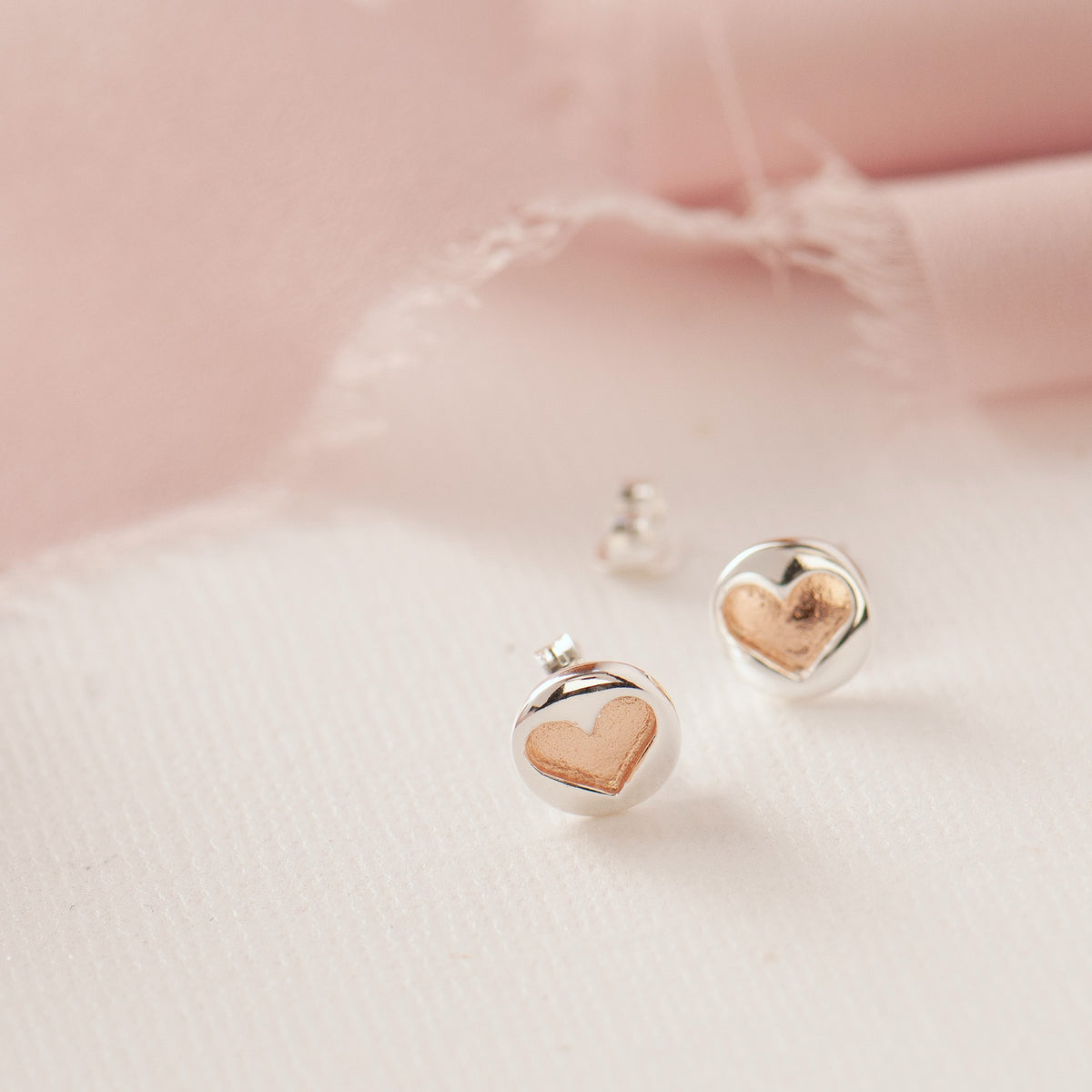A pair of silver heart stud earrings with a rose gold heart inlay, displayed on a soft pink and white textured surface.