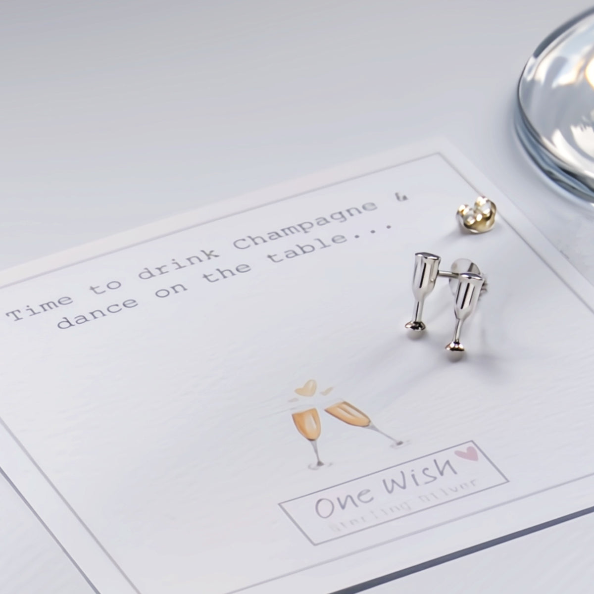 A pair of silver champagne flute stud earrings displayed on a white card that reads 'Time to drink Champagne & dance on the table.