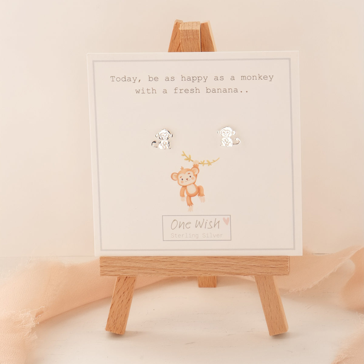 A pair of small silver monkey stud earrings displayed on a white card with a cute monkey illustration and the text 'Today, be as happy as a monkey with a fresh banana.