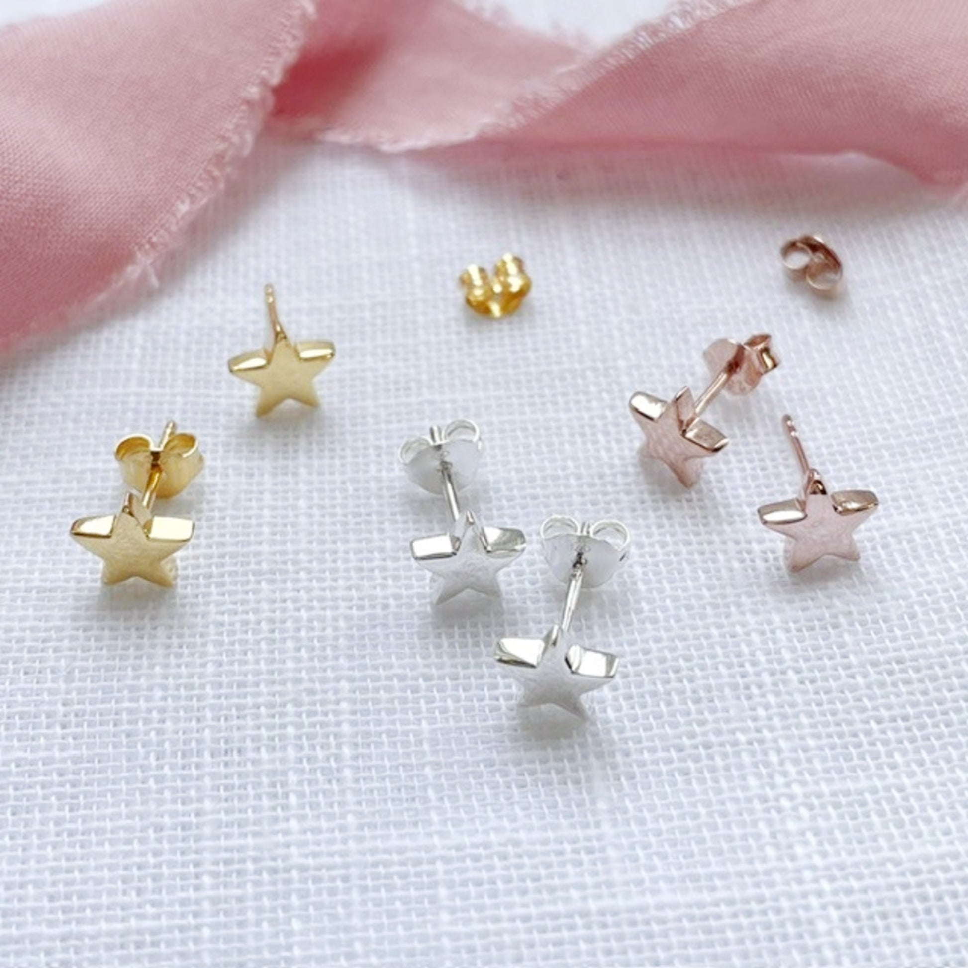 A set of small star-shaped stud earrings in gold, silver, and rose gold, arranged on a textured white fabric with a soft pink ribbon in the background.