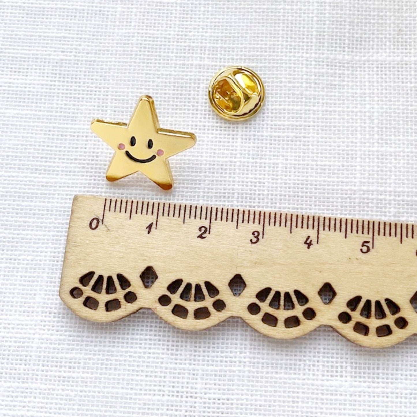 Gold star-shaped pin with a smiley face on a white fabric background with a wooden ruler for scale.