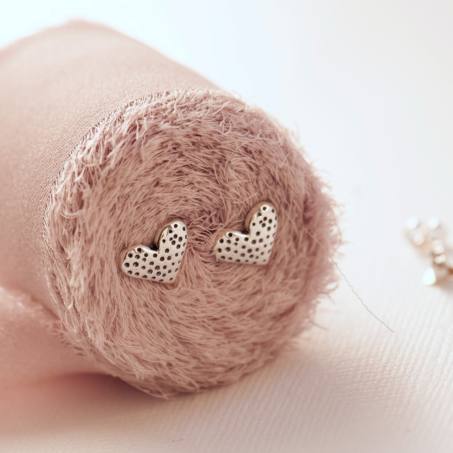 A pair of small heart-shaped stud earrings displayed on a textured, rolled piece of dusky pink fabric.