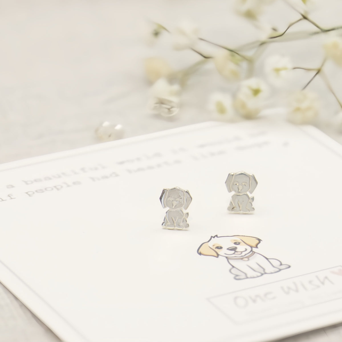 A pair of small silver-tone dog-shaped stud earrings displayed on a white card with a puppy illustration, styled with soft white flowers in the background.