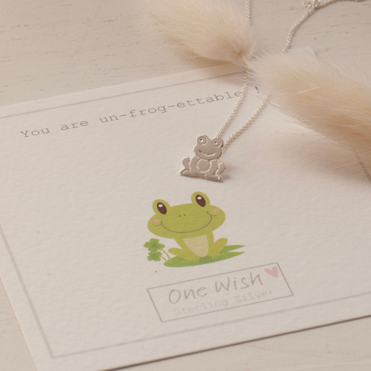 A silver necklace with a tiny frog pendant on a white card that reads 'You are Un-Frog-ettable' with pendant on the top and a signature flower envelope jewellery box.