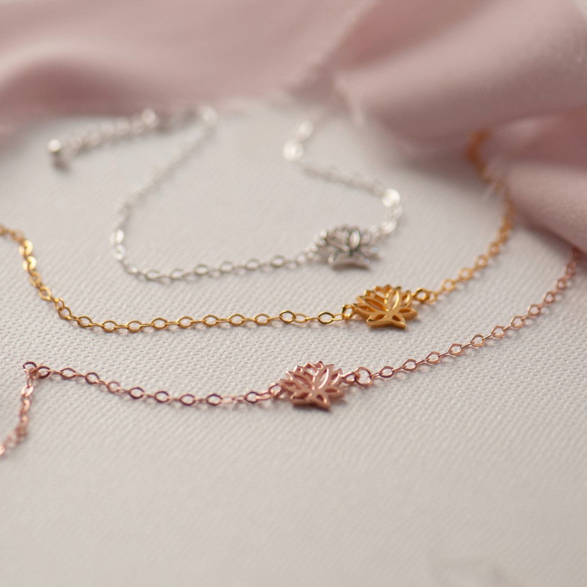 Three delicate chain necklaces with small lotus flower charms in silver, gold, and rose gold, arranged on a textured cream fabric with soft pink satin in the background.