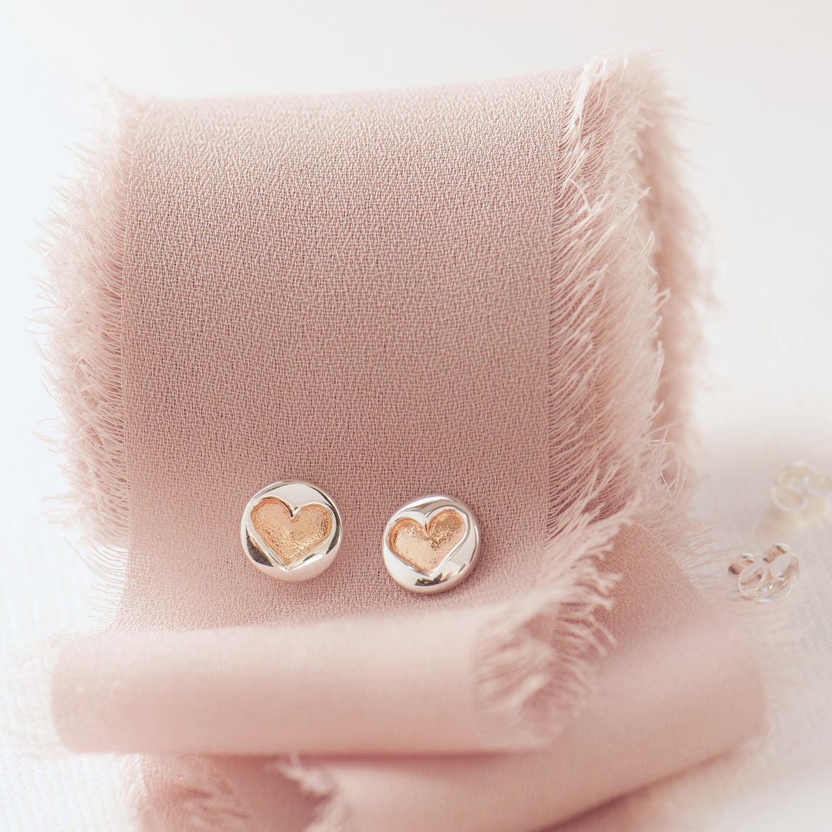 A pair of small round stud earrings with a polished silver-tone outer surface and a smaller gold-tone heart design in the center, displayed on a soft blush pink fabric stand.