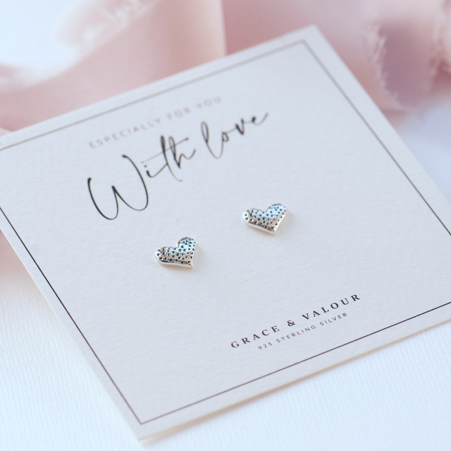 A pair of small heart-shaped stud earrings with a textured finish, displayed on a white branded card that reads 'With love' and 'Grace & Valour'.