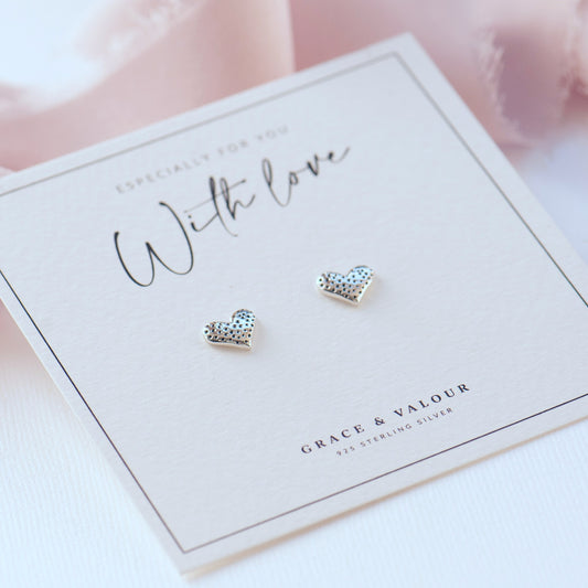 A pair of small heart-shaped stud earrings with a textured finish, displayed on a white branded card that reads 'With love' and 'Grace & Valour'.