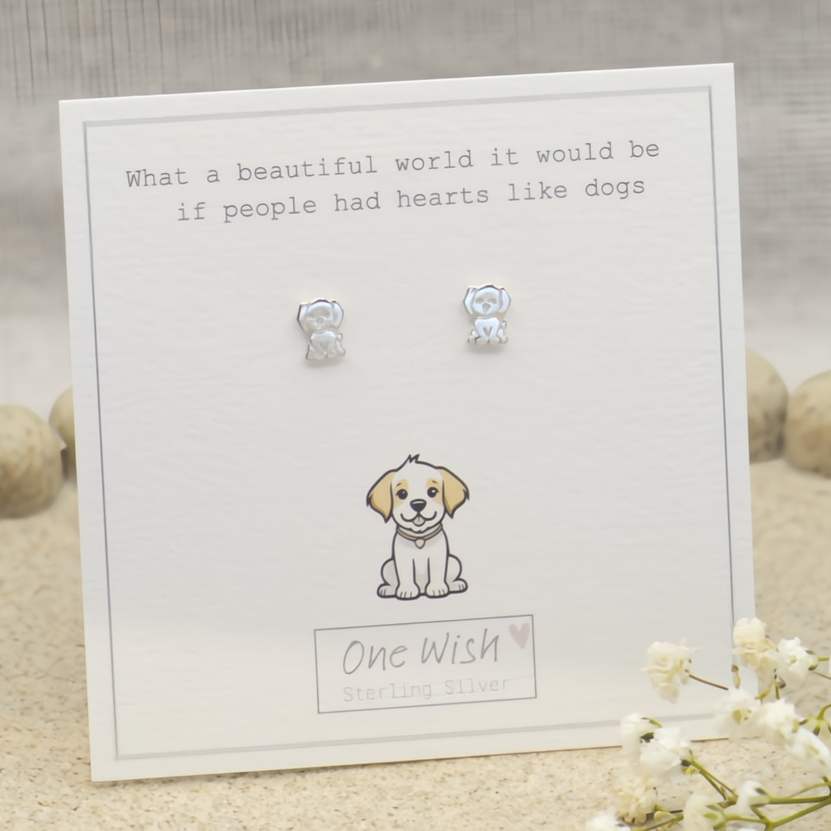 A pair of sterling silver stud earrings shaped like small dogs, displayed on a white gift card with printed text and a puppy illustration. The card is labeled 'One Wish Sterling Silver' and is photographed on a soft neutral surface with blurred decorative stones and white flowers.